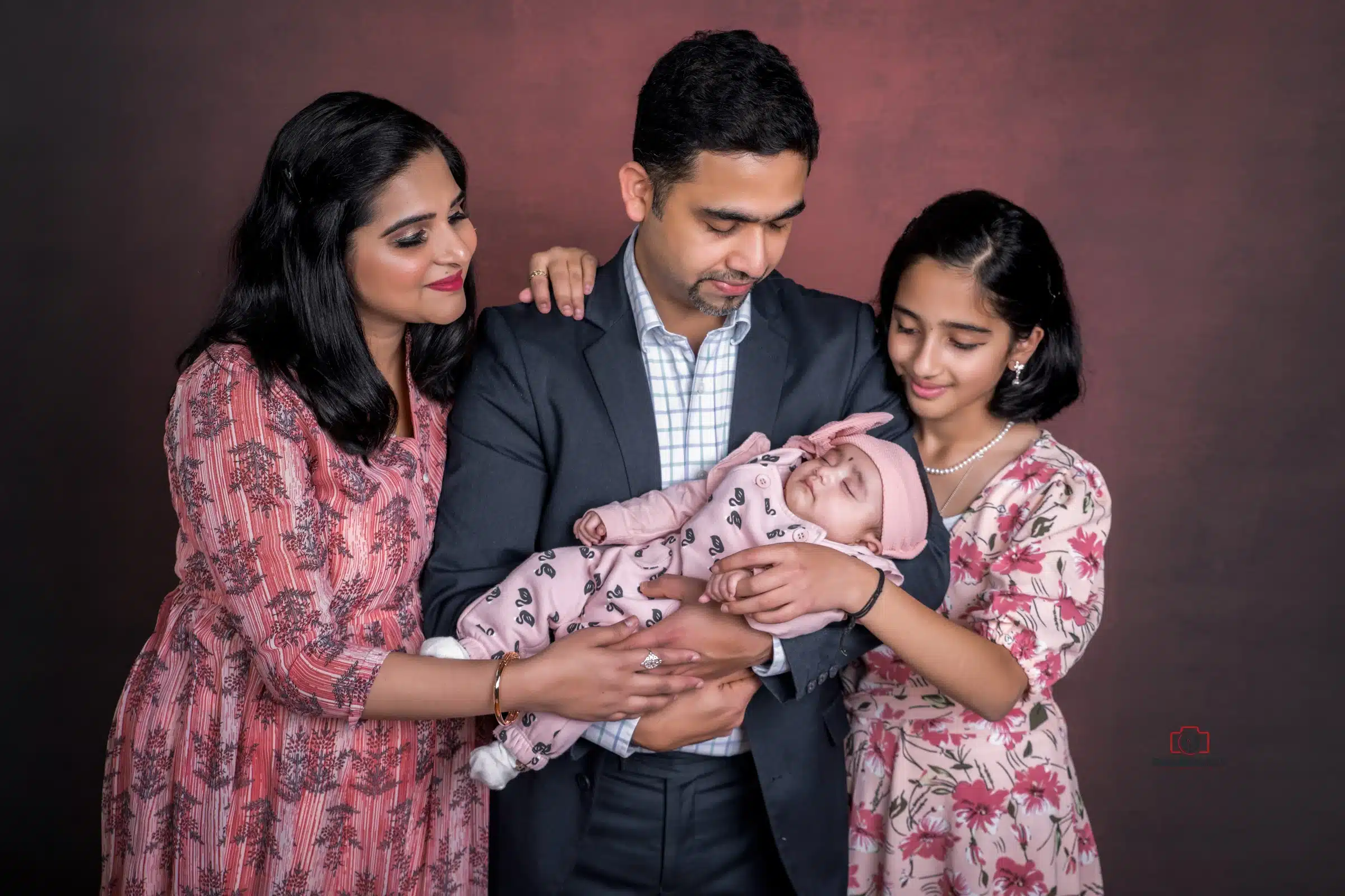 Family portrait featuring a father holding a sleeping baby in his arms, with the mother and older daughter standing beside him, all smiling warmly in coordinated pink and floral outfits.
